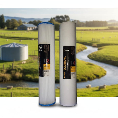 Two white cylindrical water filters with black and yellow labels, one labeled 20 micron and one labeled 1 micron, shown against a blurred pastoral farm background with a stream, green fields, and a silo.